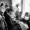 A patient seated in a clinic waiting room using a smartphone to complete a digital intake form, with reception staff in the background.