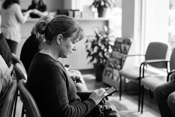A patient seated in a clinic waiting room using a smartphone to complete a digital intake form, with reception staff in the background.