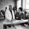 A clinician and a practice administrator reviewing patient records at a healthcare clinic reception desk, with a busy waiting room behind them.